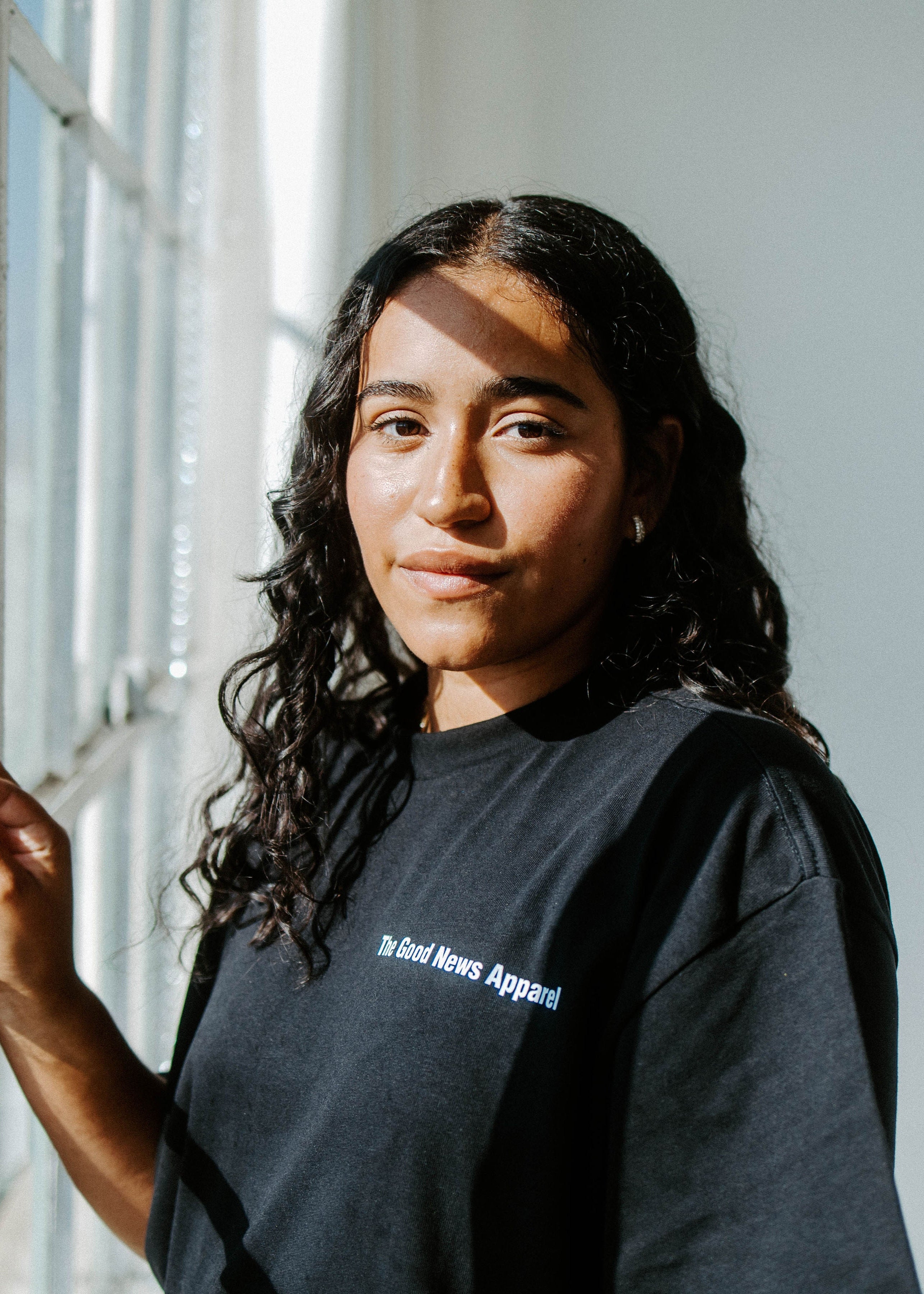 Person wearing a black t-shirt with text, standing by a window with a view of greenery.