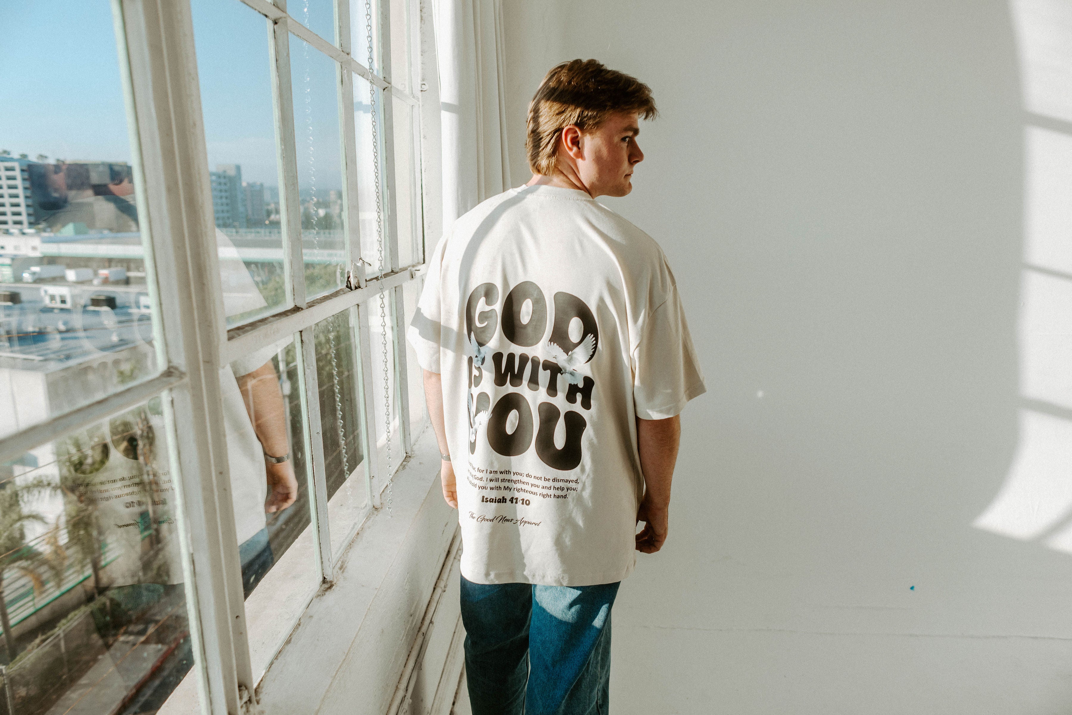 Person wearing a white t-shirt with 'God is with you' text, standing by a window with a cityscape view.