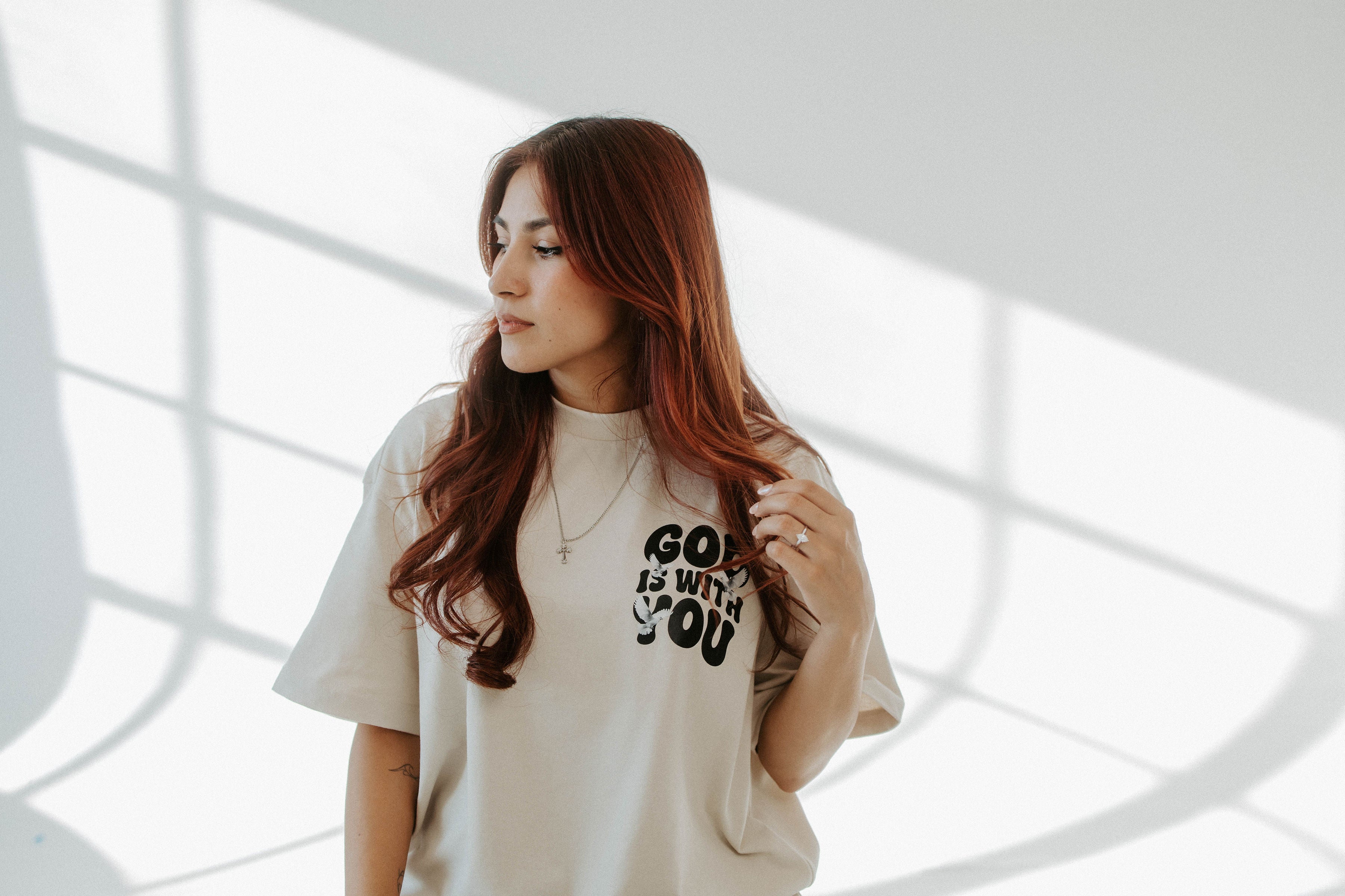 Woman wearing a white t-shirt with text, standing in a room with light filtering through.