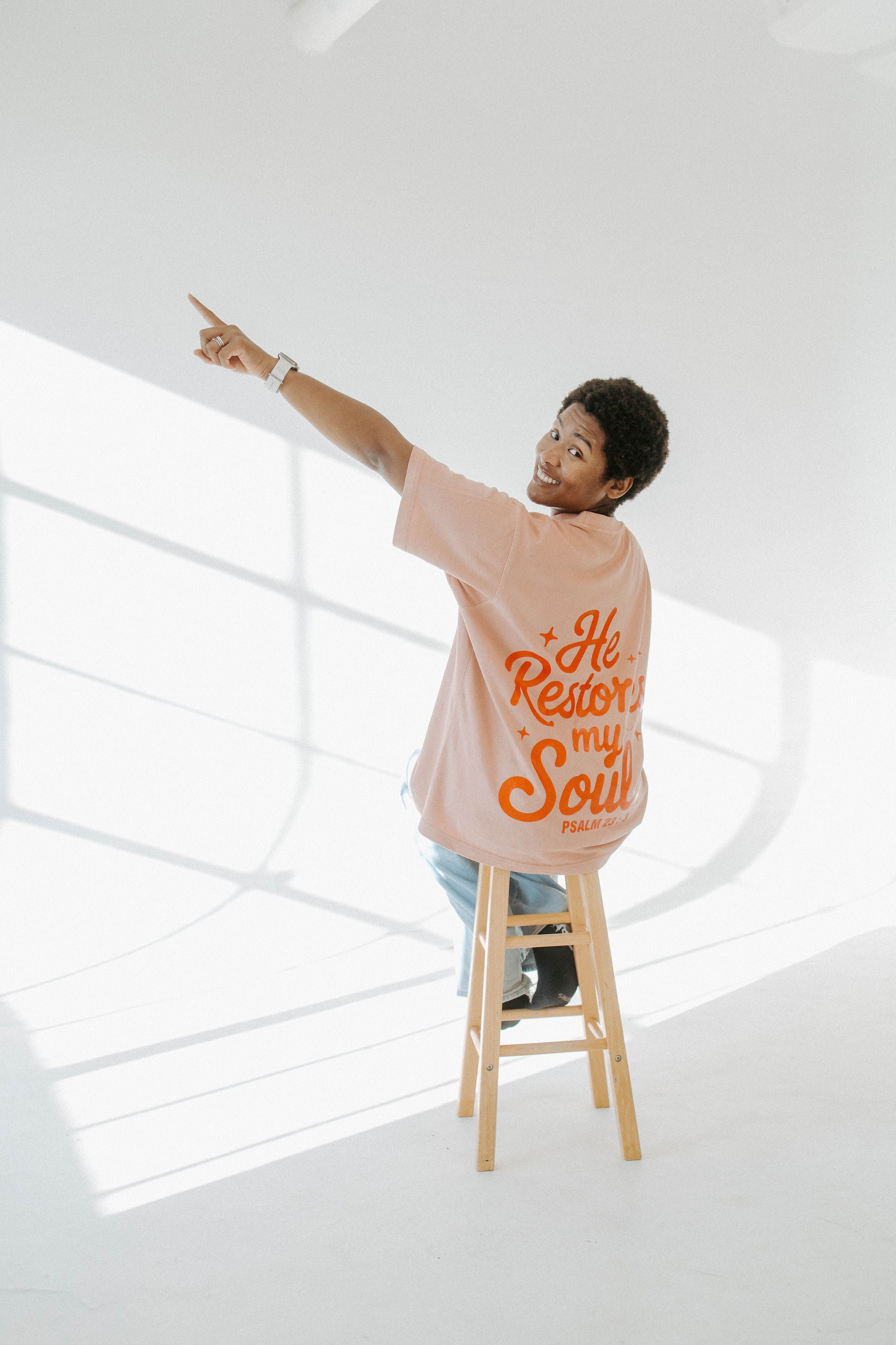 Person on a stool wearing a beige t-shirt with text, pointing upwards in a bright room.