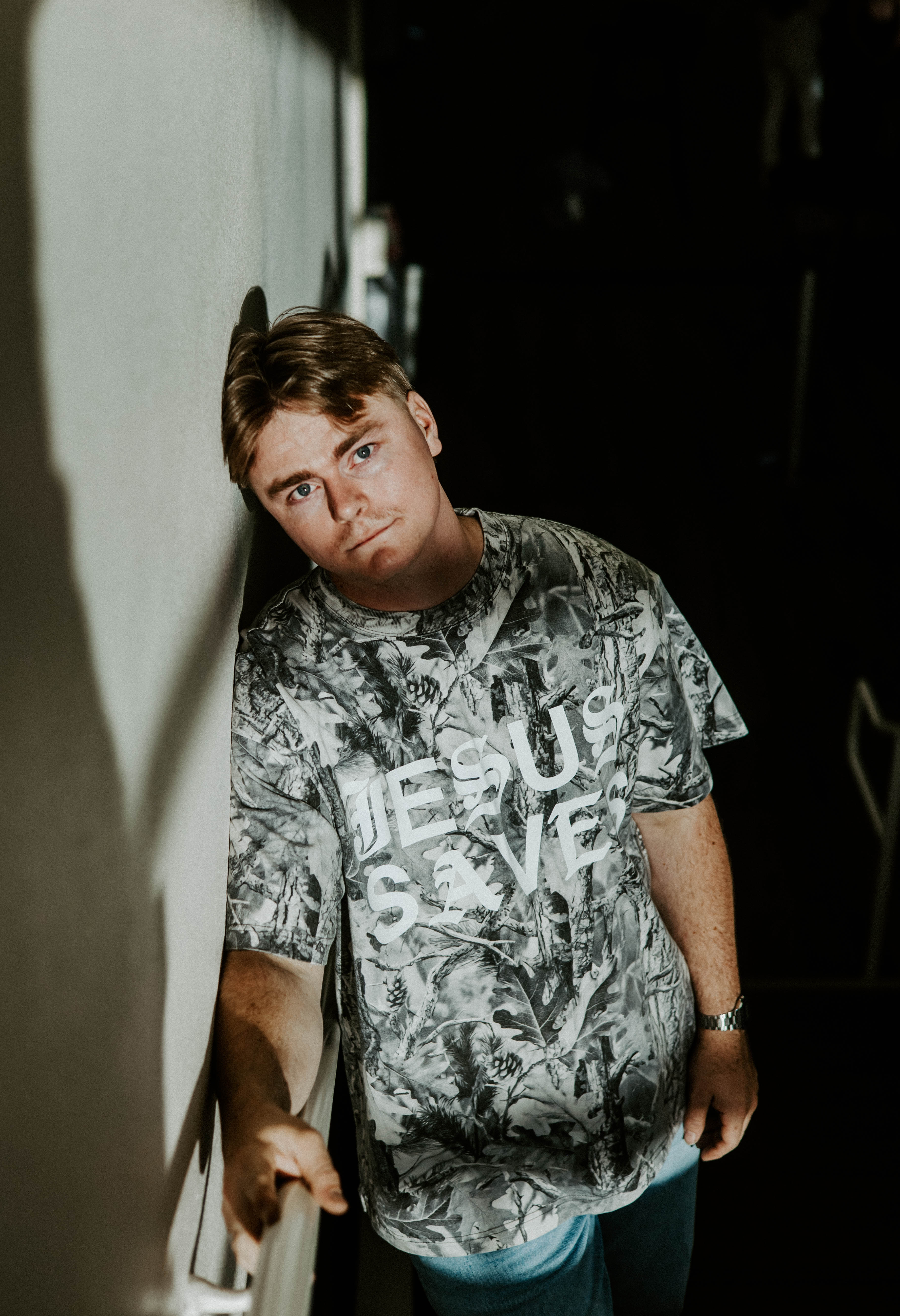 Person wearing a graphic t-shirt leaning against a wall indoors