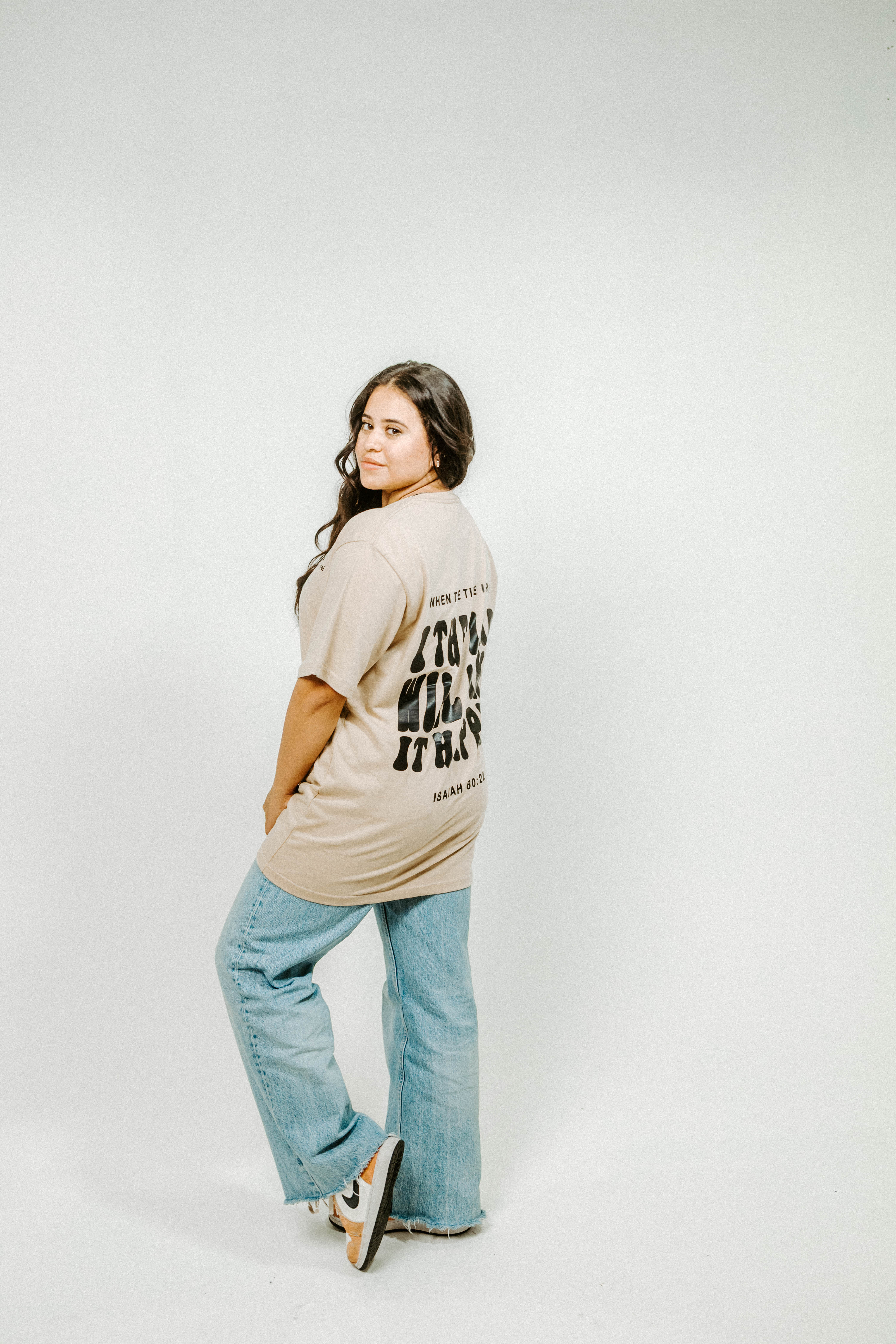 Woman wearing a beige t-shirt with text and light blue jeans on a white background