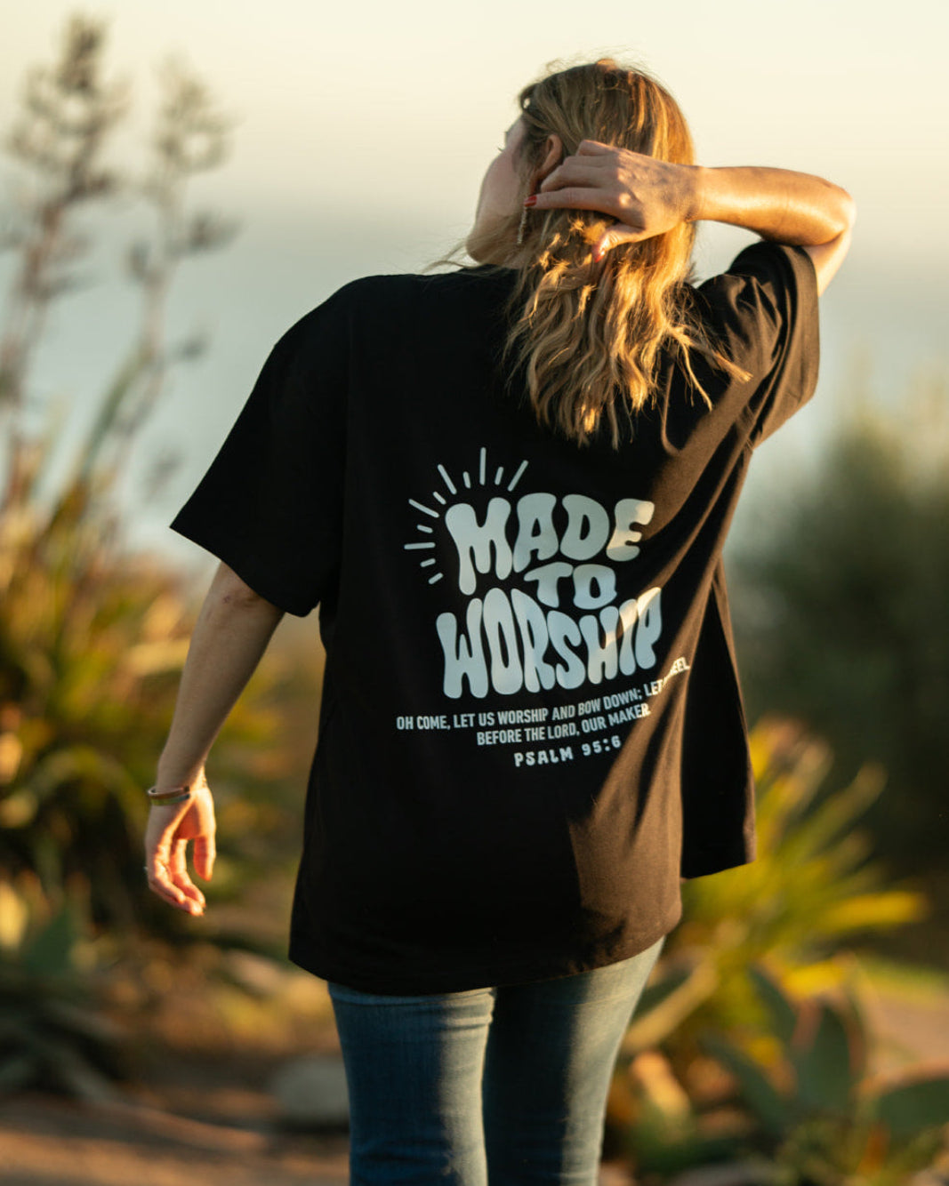 Person wearing a black t-shirt with 'Made to Worship' text, standing outdoors with plants in the background.
