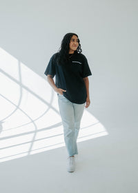 Person wearing a black t-shirt and light blue jeans standing in a modern indoor setting with large windows.