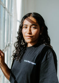 Person wearing a black t-shirt with text, standing by a window with a view of greenery.