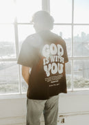 Person wearing a black t-shirt with 'God is with you' text, standing in front of large windows with a cityscape view.
