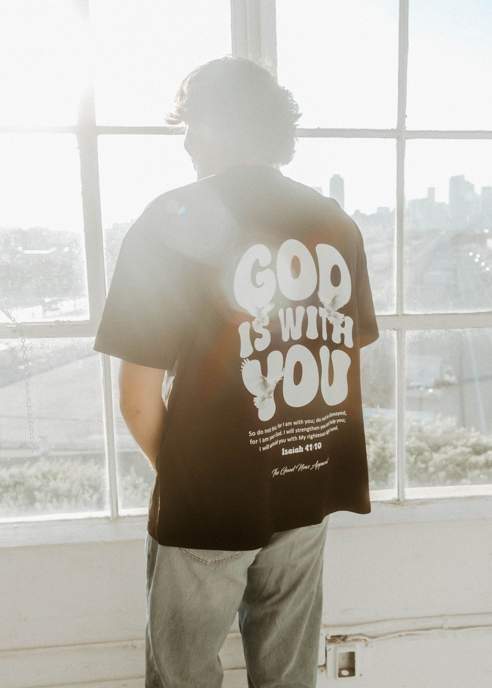 Person wearing a black t-shirt with 'God is with you' text, standing in front of large windows with a cityscape view.