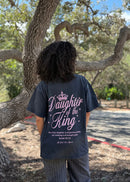 A person viewed from behind wearing a dark charcoal gray graphic t-shirt with 'Daughter of the King' printed in white and pink, standing outdoors in front of a large tree.