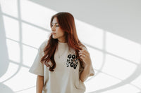 Woman wearing a white t-shirt with text, standing in a room with light filtering through.