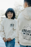 A child standing outdoors wearing a soft beige hoodie with the faith message “Created With a Purpose” printed on the back.