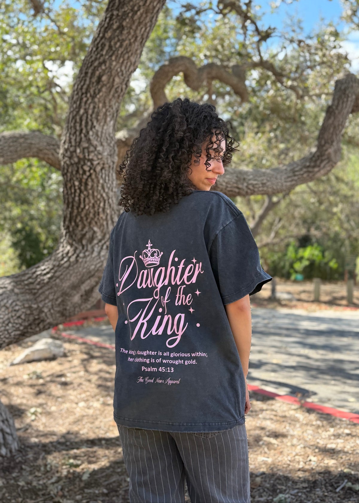A woman stands outdoors in front of a large tree, wearing a dark charcoal gray graphic t-shirt with 'Daughter of the King' and a Bible verse printed in pink and white on the back, paired with gray pinstripe pants.