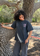 A woman with curly dark hair stands outdoors leaning against a large tree trunk, wearing a faded black graphic t-shirt with pink script text on the chest and gray pinstriped pants.