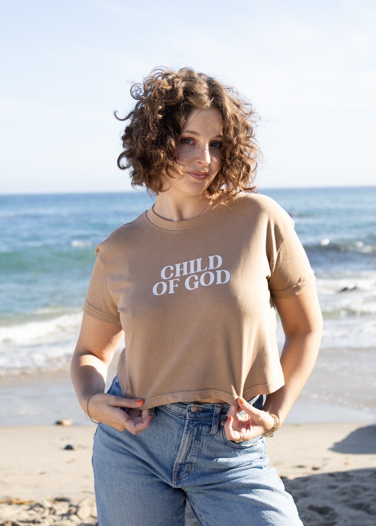 A woman with curly brown hair stands on a sandy beach wearing a beige cropped t-shirt with 'CHILD OF GOD' printed in white text, paired with light blue jeans, with the ocean and rocks in the background.