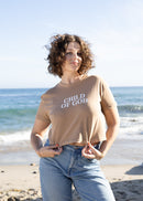 A woman with curly brown hair stands on a sandy beach wearing a beige graphic t-shirt that reads 'CHILD OF GOD' and light-wash blue jeans, with the ocean and rocks in the background.