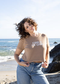 A woman with curly brown hair stands on a sandy beach wearing a beige graphic t-shirt that reads 'CHILD OF GOD' in white text, paired with light blue jeans. The background features the blue ocean, white waves, and dark rocks under a bright sky.