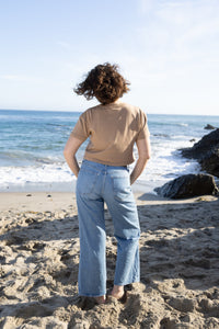 A woman stands on a sandy beach facing the ocean, wearing a beige short-sleeve t-shirt and light blue wide-leg jeans.
