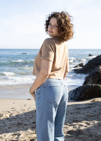 A woman with curly brown hair stands on a sandy beach looking back over her shoulder. She is wearing a beige short-sleeve crop top and light blue jeans, with the ocean, rocks, and sky in the background.