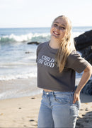 A smiling young woman stands on a beach wearing a dark gray cropped t-shirt with 'CHILD OF GOD' printed in white text across the chest, paired with light blue jeans.
