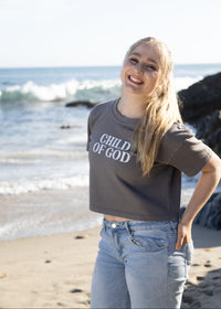 A smiling young woman stands on a beach wearing a dark gray cropped t-shirt with 'CHILD OF GOD' printed in white text across the chest, paired with light blue jeans.