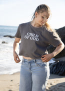 A woman standing on a beach wearing a dark gray cropped t-shirt with the text 'CHILD OF GOD' in white, paired with light blue jeans.