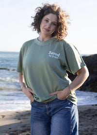 A woman with curly brown hair stands on a sandy beach wearing an olive green t-shirt with 'Saved by Chris' printed in white on the left chest, paired with blue jeans.