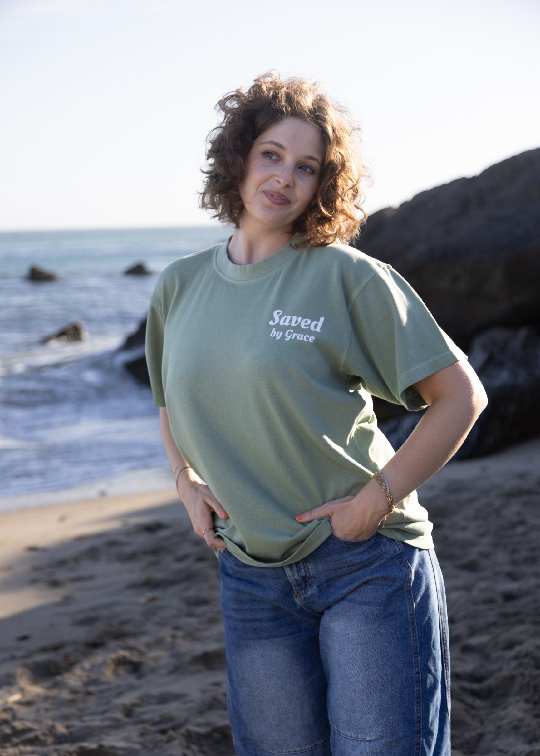 A woman with curly brown hair stands on a sandy beach wearing an olive green t-shirt with the text 'Saved by Grace' in white on the chest, paired with blue jeans.