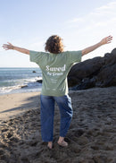 A person standing on a sandy beach with arms outstretched, wearing a light green T-shirt with 'Saved by Green' printed in white on the back, paired with blue jeans.