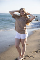A woman stands on a sandy beach with the ocean in the background, wearing a tan short-sleeve t-shirt and white shorts.