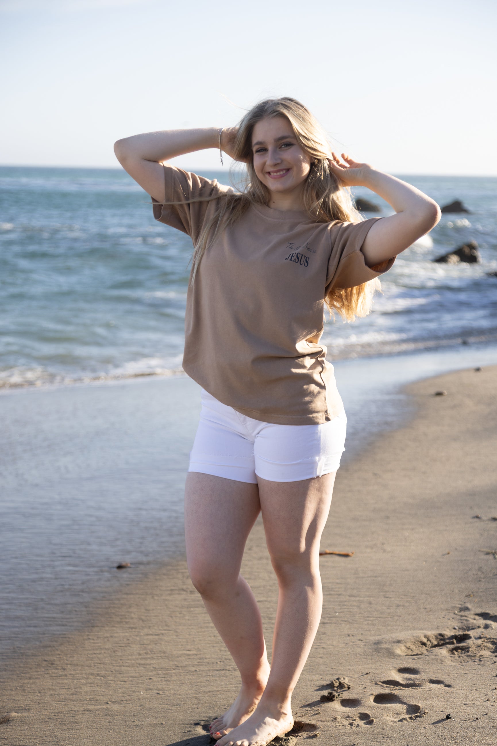 A woman stands on a sandy beach with the ocean in the background, wearing a tan short-sleeve t-shirt and white shorts.