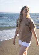 A woman walking on a sandy beach wearing a brown t-shirt with 'Jesus' printed on the chest and white shorts, with the ocean in the background.