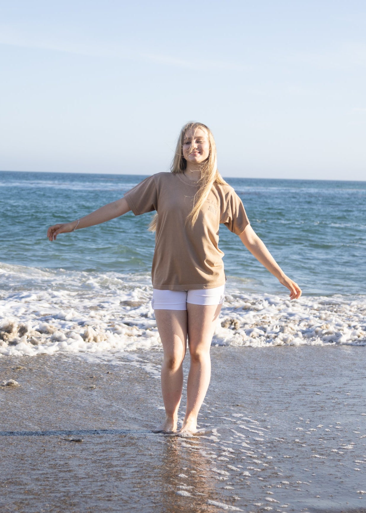 A woman stands barefoot in shallow ocean water on a beach, wearing a loose beige t-shirt and white shorts, with her arms outstretched. The sea and sky are visible in the background.