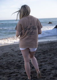 A woman stands on a sandy beach facing the ocean, wearing a beige graphic t-shirt with 'The Good News Club' printed in black script on the back, paired with white shorts.