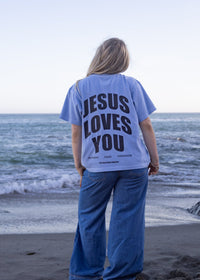 A woman stands on a beach facing the ocean, wearing a light blue graphic t-shirt with the words 'JESUS LOVES YOU' printed in black on the back, paired with wide-leg blue jeans.