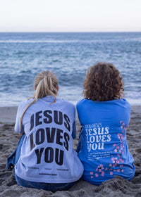 Two people sitting on a sandy beach facing the ocean, wearing matching graphic t-shirts with the text 'JESUS LOVES YOU' printed on the back.