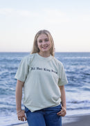 A young woman stands on a beach wearing a light sage green graphic t-shirt with the text 'All Hail King Jesus' printed in black on the chest, paired with blue jeans.