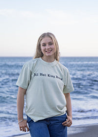 A young woman stands on a beach wearing a light sage green graphic t-shirt with the text 'All Hail King Jesus' printed in black on the chest, paired with blue jeans.