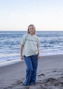 A woman stands on a sandy beach with the ocean and rocks behind her, wearing a light sage green graphic T-shirt and blue jeans.