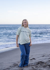 A woman stands on a sandy beach with the ocean and rocks behind her, wearing a light sage green graphic T-shirt and blue jeans.