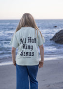 A woman standing on a beach with her back to the camera, wearing a light sage green t-shirt with 'All Hail King Jesus' printed in black gothic font on the back, paired with blue jeans.
