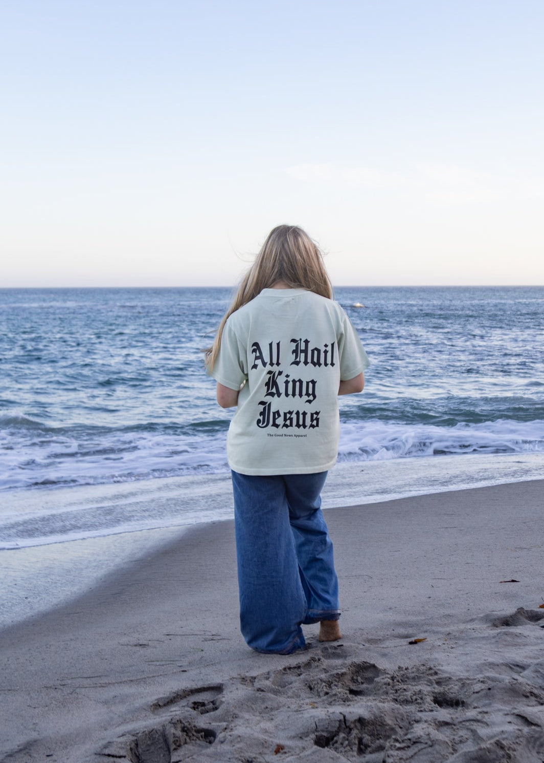 A woman stands on a sandy beach facing the ocean, wearing a cream-colored t-shirt with 'All Hail The King Jesus' printed in black on the back, paired with blue jeans.