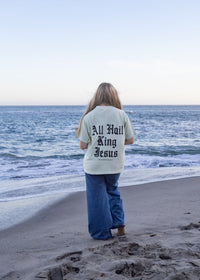 A woman stands on a sandy beach facing the ocean, wearing a cream-colored t-shirt with 'All Hail The King Jesus' printed in black on the back, paired with blue jeans.