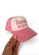 Pink and white cap with 'Saved by Grace' text held by a hand on a white background