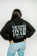 Person wearing a black hoodie with 'Victory Over Fear' text on the back, standing against a white background.