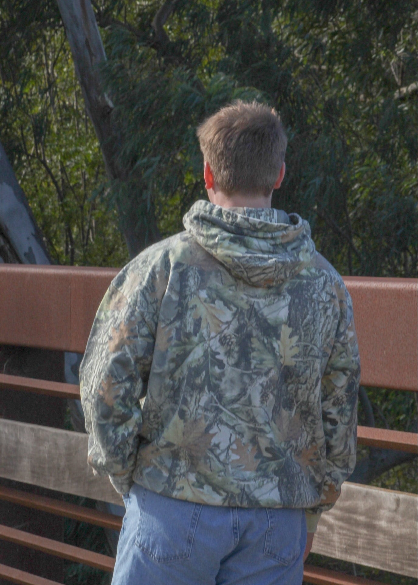 Person wearing a camouflage jacket and blue pants standing on a wooden bridge with trees in the background