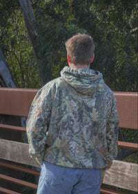 Person wearing a camouflage jacket and blue pants standing on a wooden bridge with trees in the background