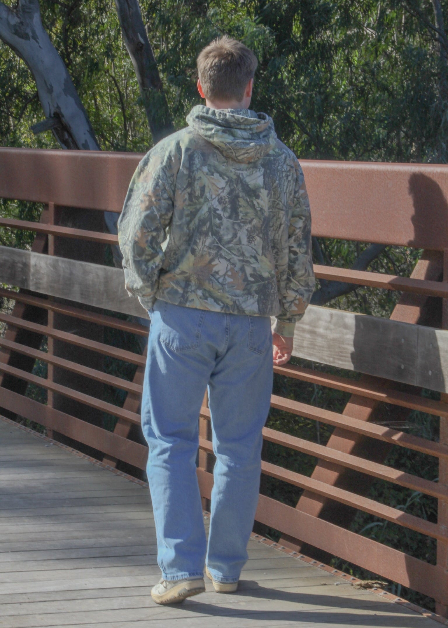 Person wearing a camouflage jacket and blue jeans standing on a wooden bridge with trees in the background