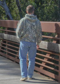 Person wearing a camouflage jacket and blue jeans standing on a wooden bridge with trees in the background