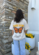 Person wearing a white t-shirt with a butterfly graphic and text, standing in front of a stone wall with potted plants.