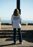 Person wearing a white t-shirt with 'Saved by Grace' text, standing on steps with ocean view.