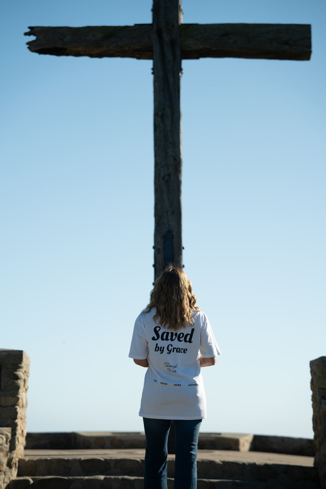 Person wearing a 'Saved by Grace' shirt standing in front of a cross against a clear blue sky.